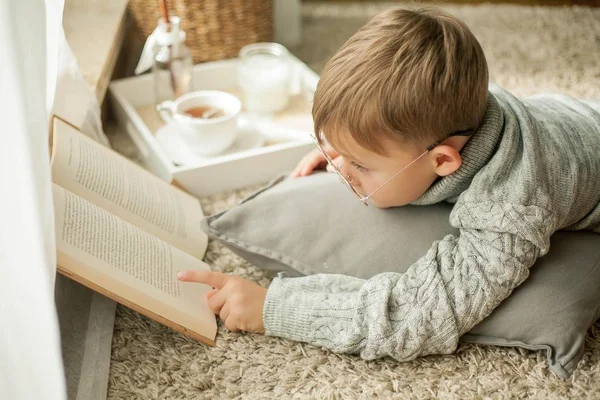 A beautiful little boy in a knitted sweater is reading by the window with a mug of hot tea. Cozy. Autumn. — Stock Photo, Image