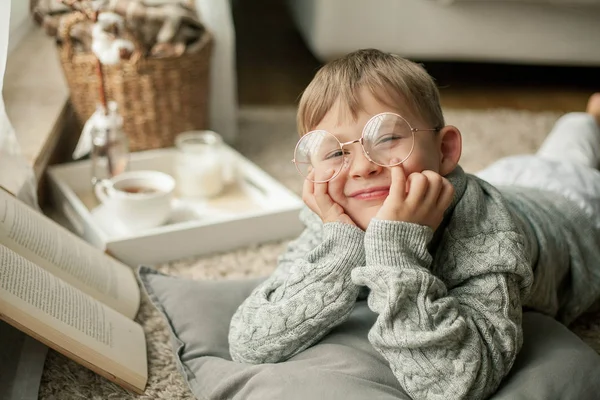 Een mooie kleine jongen in een gebreide trui is lezen bij het raam met een mok warme thee. Gezellige. Herfst. — Stockfoto