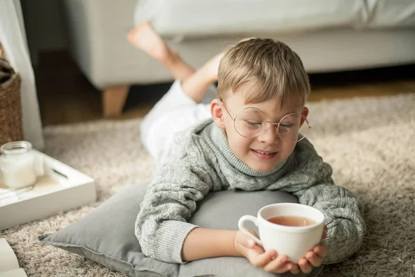 En vacker liten pojke i en stickad tröja läser av fönstret med en mugg hett te. Mysiga. Hösten. — Stockfoto