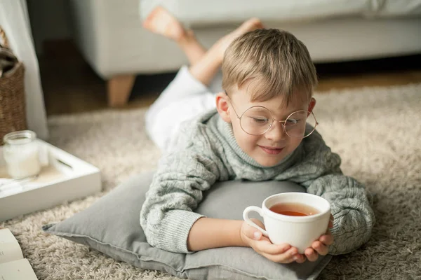 Um menino bonito em uma camisola de malha está lendo pela janela com uma caneca de chá quente. Aconchegante. Outono . — Fotografia de Stock