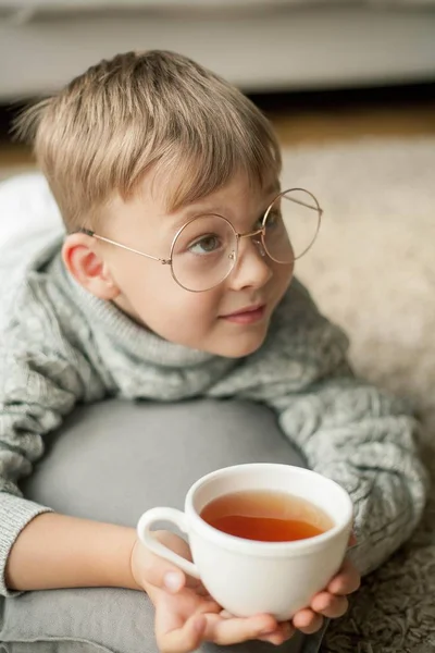 Um menino bonito em uma camisola de malha está lendo pela janela com uma caneca de chá quente. Aconchegante. Outono . — Fotografia de Stock