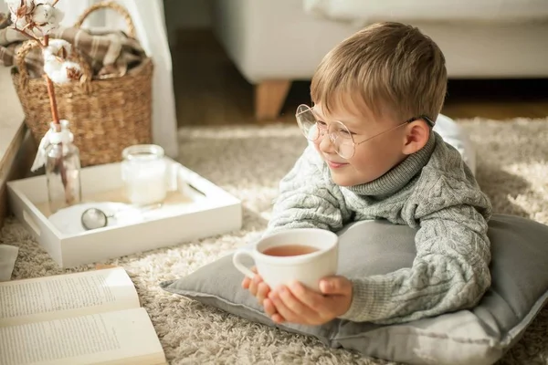 Um menino bonito em uma camisola de malha está lendo pela janela com uma caneca de chá quente. Aconchegante. Outono . — Fotografia de Stock