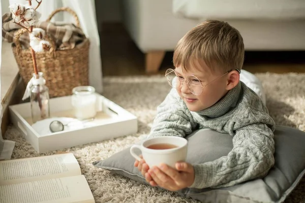 En vacker liten pojke i en stickad tröja läser av fönstret med en mugg hett te. Mysiga. Hösten. — Stockfoto