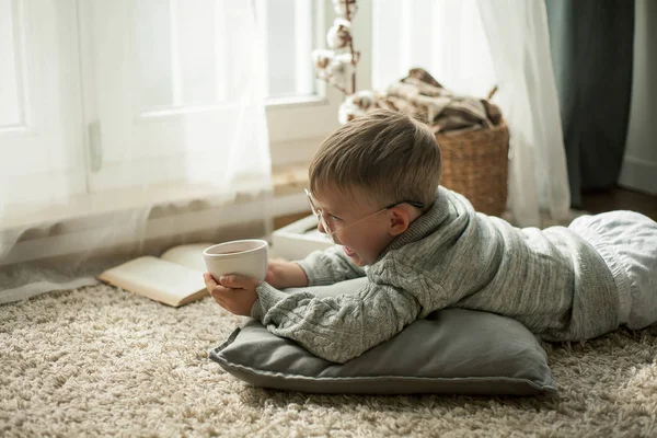 Un bel bambino con un maglione a maglia sta leggendo vicino alla finestra con una tazza di tè caldo. Accogliente. Autunno . — Foto Stock