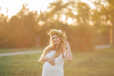Güneşte kalmış hamile bir kadının portresi. Sahada başında çelenk olan güzel, hamile bir kadın. Annelik. Sıcak sonbahar.