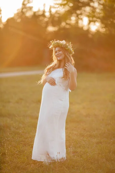 Retrato de una mujer embarazada bajo el sol. Joven mujer embarazada hermosa con una corona en la cabeza en el campo. Maternidad. Otoño cálido . — Foto de Stock