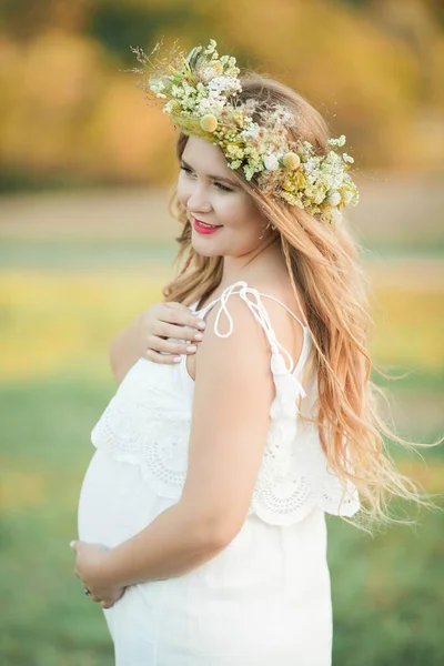 太陽の下で妊婦の肖像画。フィールドに彼女の頭の上に花輪を持つ若い美しい妊婦。母親。暖かい秋. — ストック写真