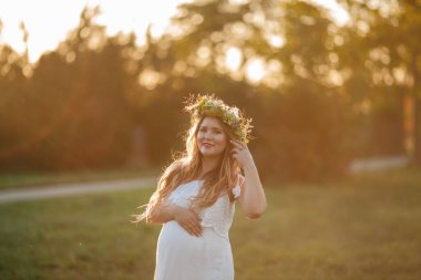 Güneşte kalmış hamile bir kadının portresi. Sahada başında çelenk olan güzel, hamile bir kadın. Annelik. Sıcak sonbahar.