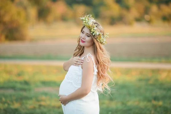 Πορτρέτο μιας εγκύου στον ήλιο. Μια νεαρή όμορφη έγκυος γυναίκα με ένα στεφάνι στο κεφάλι της στο χωράφι. Μητρότητα. Ζεστό φθινόπωρο. — Φωτογραφία Αρχείου
