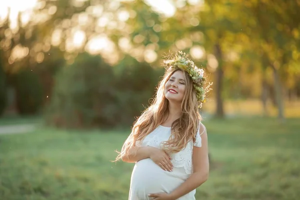 太陽の下で妊婦の肖像画。フィールドに彼女の頭の上に花輪を持つ若い美しい妊婦。母親。暖かい秋. — ストック写真