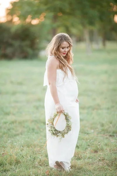 Retrato de una mujer embarazada bajo el sol. Joven mujer embarazada hermosa con una corona en la cabeza en el campo. Maternidad. Otoño cálido . — Foto de Stock