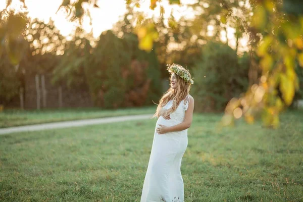 太陽の下で妊婦の肖像画。フィールドに彼女の頭の上に花輪を持つ若い美しい妊婦。母親。暖かい秋. — ストック写真