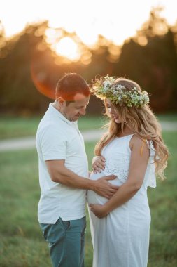 Bebek bekleyen genç ve güzel bir çift. Kafasında çelenk olan güzel hamile bir kadın karnına tutunuyor. Annelik. Sıcak sonbahar.