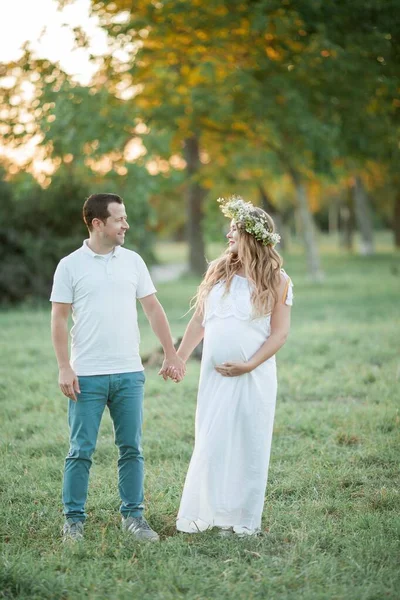 Jovem casal lindo esperando um bebê. Jovem mulher grávida bonita com uma coroa de flores na cabeça segura uma barriga. Maternidade. Outono quente . — Fotografia de Stock