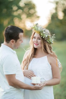 Bebek bekleyen genç ve güzel bir çift. Kafasında çelenk olan güzel hamile bir kadın karnına tutunuyor. Annelik. Sıcak sonbahar.