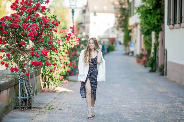 Young beautiful woman walks in a European city. Portrait of a young woman near blooming roses. Europe. — Stock Photo, Image