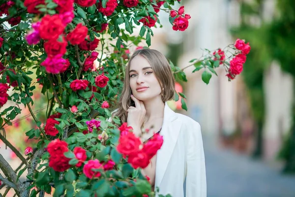Nuori kaunis nainen kävelee eurooppalaisessa kaupungissa. Nuoren naisen muotokuva lähellä kukkivia ruusuja. Eurooppa. — kuvapankkivalokuva