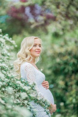 Dantelli, beyaz elbiseli güzel hamile bir kadın çiçek parkında yürüyor. Güzel hamile bir sarışının portresi..