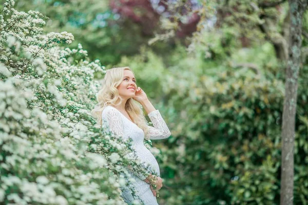 穿着花边白色连衣裙的年轻漂亮的孕妇走在一个开花的公园里 美丽的怀孕金发美女的画像 — 图库照片