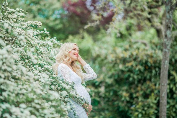 Young beautiful pregnant woman in a lace white dress walks in a flowering park. Portrait of a beautiful pregnant blonde.
