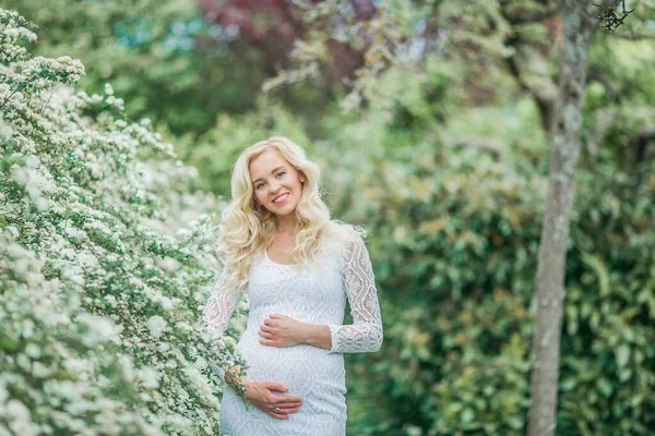 レースの白いドレスを着た若い美しい妊婦が開花公園を歩いています 美しいです妊娠中のブロンドの肖像 — ストック写真