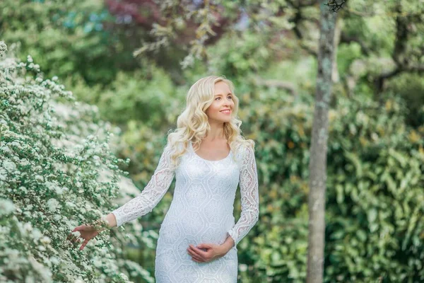 穿着花边白色连衣裙的年轻漂亮的孕妇走在一个开花的公园里 美丽的怀孕金发美女的画像 — 图库照片