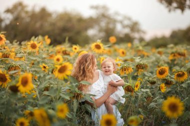 Genç ve güzel bir anne küçük kıvırcık bir kız çocuğu tutuyor. Annelik. Kıvırcık anne ve küçük kızı ayçiçeği tarlasında.