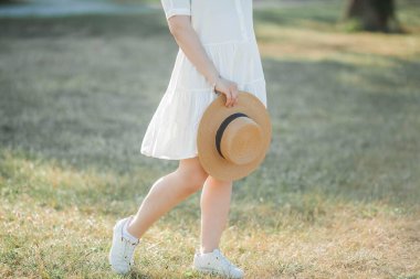 Güneş ışınlarındaki genç kadın portresi. Hasır şapkalı beyaz elbiseli güzel bir kadın parkta yürüyor..