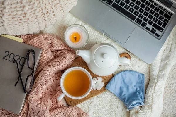 Laptop, face mask, hot tea mug, candle, glasses, kettle, wooden stand. Quarantine. Home Office. Cozy breakfast.