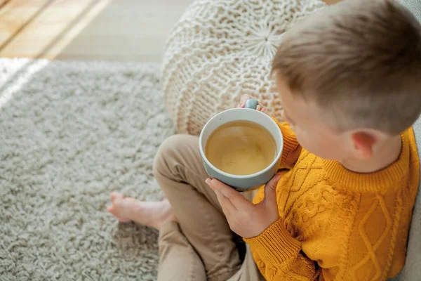 オレンジのセーターに身を包んだ可愛い男の子がお茶を飲んでいる 男の子の居心地の良い肖像画家に座って — ストック写真