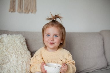 2 yaşında, rahat örgü süveter giyen tatlı küçük bir kız kanepede kahvaltı yapıyor. Samimi.