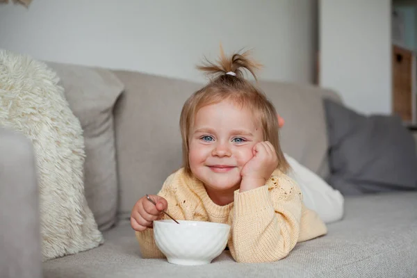 居心地の良いニットセーターのかわいい女の子2歳はソファの上で朝食を取っています 居心地の良い — ストック写真