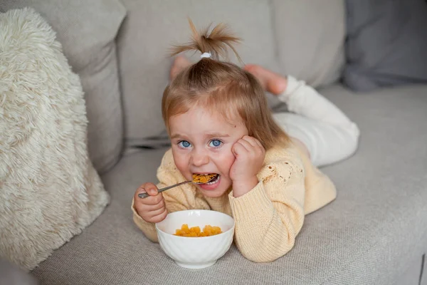 Een schattig klein meisje van 2 jaar oud in een gezellig gebreide trui zit op de bank te ontbijten. Gezellig. Vallen.. — Stockfoto