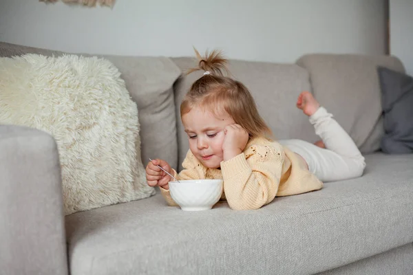 Uma menina bonita de 2 anos de idade em uma camisola de malha aconchegante está tomando café da manhã no sofá. Aconchegante. Queda. — Fotografia de Stock