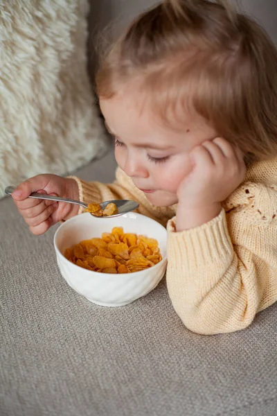Een schattig klein meisje van 2 jaar oud in een gezellig gebreide trui zit op de bank te ontbijten. Gezellig. Vallen.. — Stockfoto