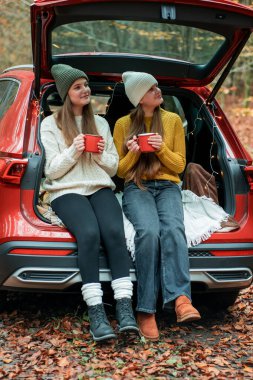 Genç kız kardeşler sonbahar ormanlarında kırmızı bir arabanın bagajında büyük bir termostan sıcak çay içiyorlar. Piknik.