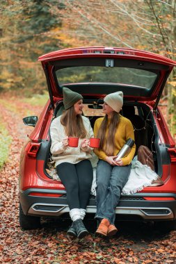 Genç kız kardeşler sonbahar ormanlarında kırmızı bir arabanın bagajında büyük bir termostan sıcak çay içiyorlar. Piknik.