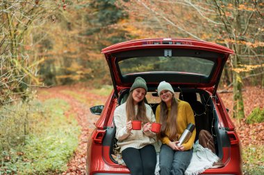 Genç kız kardeşler sonbahar ormanlarında kırmızı bir arabanın bagajında büyük bir termostan sıcak çay içiyorlar. Piknik.