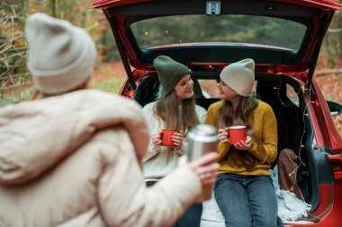 Genç anne ve genç kızları sonbahar ormanlarında kırmızı bir arabanın bagajında büyük bir termostan sıcak çay içiyorlar..