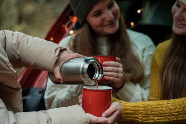 Genç anne ve genç kızları sonbahar ormanlarında kırmızı bir arabanın bagajında büyük bir termostan sıcak çay içiyorlar..