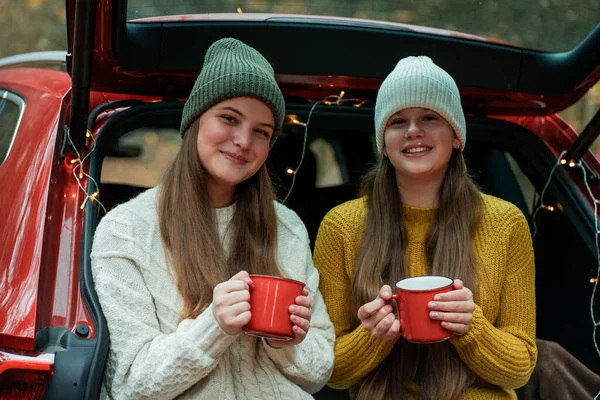 Genç kız kardeşler sonbahar ormanlarında kırmızı bir arabanın bagajında büyük bir termostan sıcak çay içiyorlar. Piknik.
