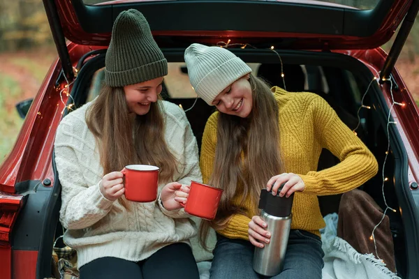 Genç kız kardeşler sonbahar ormanlarında kırmızı bir arabanın bagajında büyük bir termostan sıcak çay içiyorlar. Piknik.
