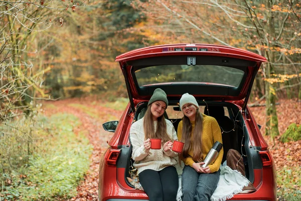 Genç kız kardeşler sonbahar ormanlarında kırmızı bir arabanın bagajında büyük bir termostan sıcak çay içiyorlar. Piknik.