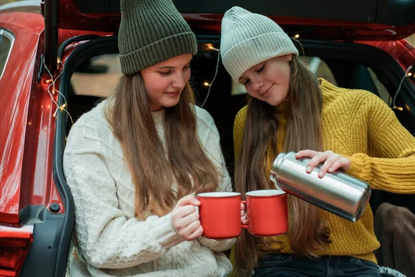 Genç kız kardeşler sonbahar ormanlarında kırmızı bir arabanın bagajında büyük bir termostan sıcak çay içiyorlar. Piknik.
