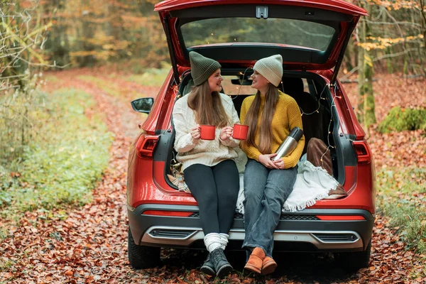 Genç kız kardeşler sonbahar ormanlarında kırmızı bir arabanın bagajında büyük bir termostan sıcak çay içiyorlar. Piknik.