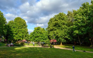 Beckenham (Büyük Londra), Kent / UK. Ağaçlı, çiçekli ve otlu Beckenham Green. İnsanlar banklarda oturuyor ve bir çocuk köpekle koşuyor. Beckenham Green, Beckenham 'ın merkezinde bir park..
