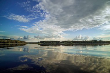 Mavi gökyüzü ve bulutlarla kaplı nehir manzarası bir yaz günü suya yansıdı. Orman ufukta görünüyor..