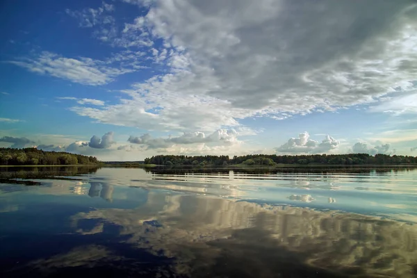 Mavi gökyüzü ve bulutlarla kaplı nehir manzarası bir yaz günü suya yansıdı. Orman ufukta görünüyor..