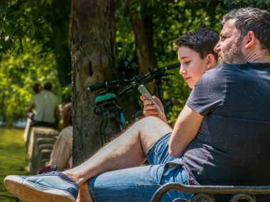 Bükreş / Romanya - 05.30.2020: Parktaki bankta oturan ve telefonuna bakan babasıyla genç bir çocuk. Akıllı telefon bağımlılığı kavramı.