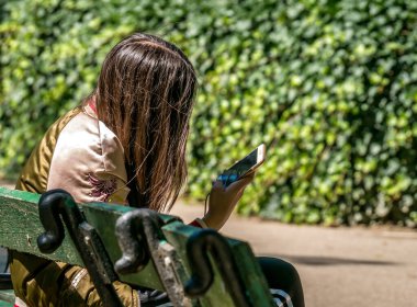 Bükreş / Romanya - 05.30.2020: Parktaki bankta oturan ve telefonuna bakan kadın. Akıllı telefon bağımlılığı kavramı.